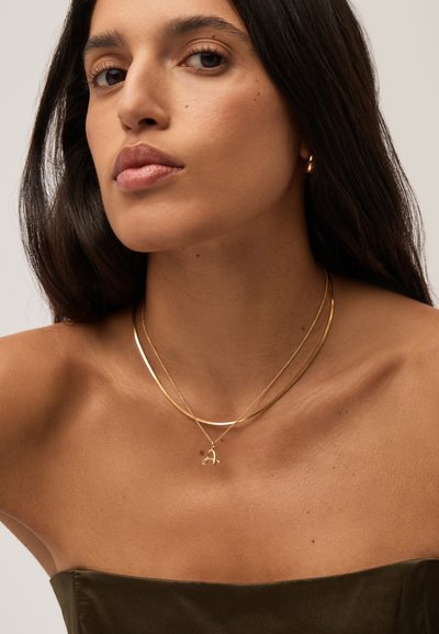 Νέα γυναίκα με μακριά σκούρα μαλλιά, που φοράει σκούρο τοπ χωρίς τιράντες, χρυσούς κρίκους στα σκουλαρίκια και δύο χρυσά κολιέ, ένα με μενταγιόν σε σχήμα "Α".