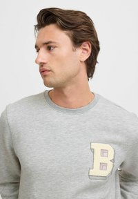 Sudadera gris de mezcla de algodón, con una letra 'B' en relieve bordada en crema y púrpura. Cuello redondo clásico y mangas largas.