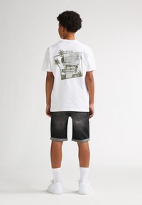 Wit katoenen T-shirt met groene grafische print met palmbomen op de achterkant; gecombineerd met donkere denim shorts en witte sneakers.