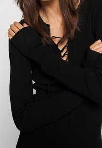 Svart ribbad stickad top med en djup V-ringning med snörningsdetalj och långa ärmar med tumhål, vilket ger en åtsittande silhuett.