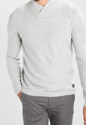 Mann trägt einen hellgrauen V-Ausschnitt-Pullover mit gerippten Bündchen und Saum, kombiniert mit dunkelgrauen Hosen, die Hände teilweise in den Taschen.