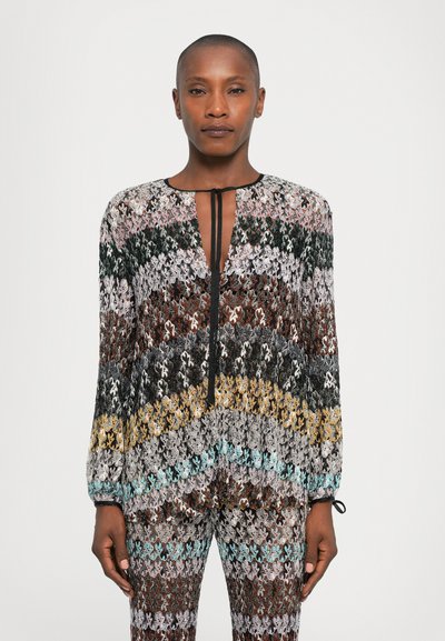 Femme portant un haut en tricot multicolore à manches longues et un pantalon assorti avec un motif géométrique, se tenant devant un fond blanc uni.