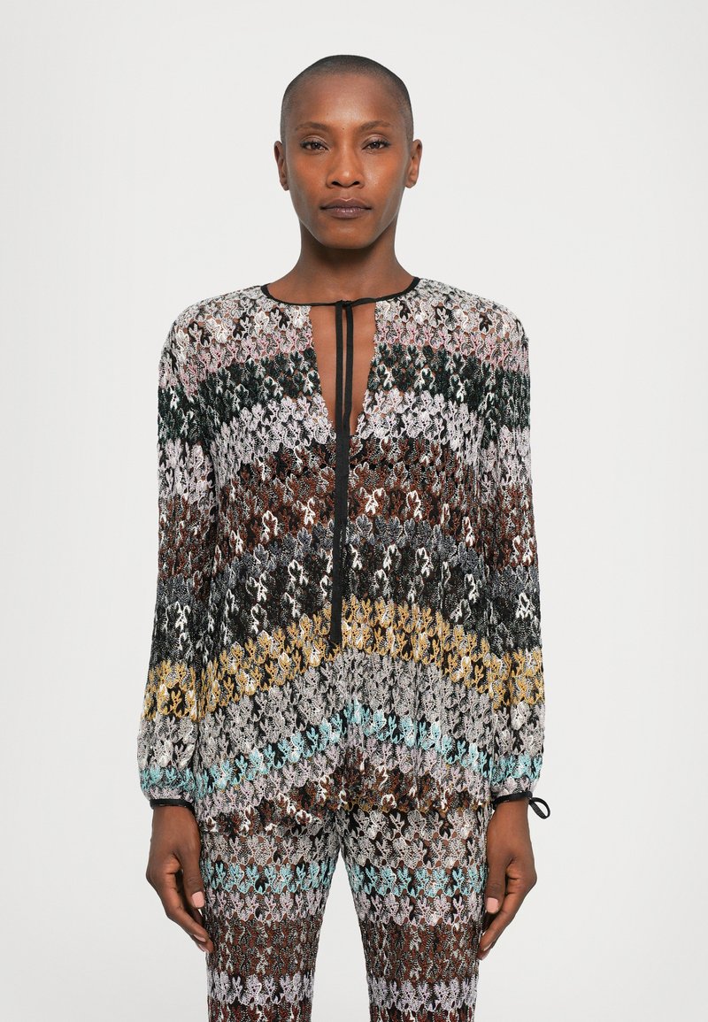 Woman wearing long-sleeve multicolored knit top and matching pants with geometric pattern, standing against a plain white background.