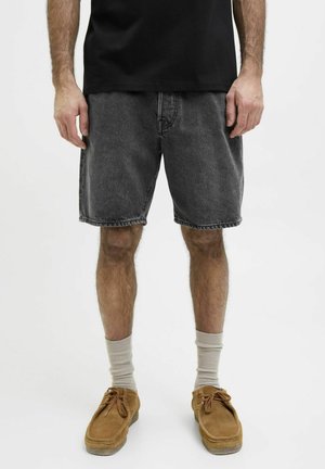 Camiseta negra de algodón combinada con pantalones cortos de mezclilla gris oscuro. Los pantalones cortos tienen un corte holgado, dobladillo deshilachado y bolsillos básicos. Zapatos de ante color arena y calcetines crema.