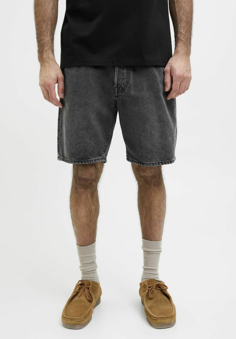 Camiseta negra de algodón combinada con pantalones cortos de mezclilla gris oscuro. Los pantalones cortos tienen un corte holgado, dobladillo deshilachado y bolsillos básicos. Zapatos de ante color arena y calcetines crema.