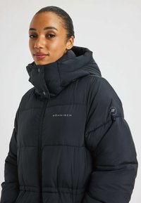 Schwarze, puffy Jacke mit hohem Kragen, ausgestattet mit einem Reißverschluss und Druckknöpfen. Mit Logodetails am linken Ärmel. Glatte Textur.