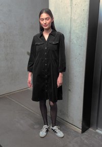 Femme portant une robe noire boutonnée, des collants noirs et des baskets blanches, debout à côté d'un mur en béton et d'une porte d'ascenseur.