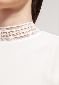 Biała bluzka z wysokim kołnierzykiem, ozdobiona koronkowym wykończeniem w trójkątne wzory, o gładkiej teksturze i minimalistycznym designie.