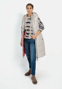 Lys grå polstret vest med hætte, der har en blød inderforing og røde detaljer, båret over en mønstret striktrøje og blå jeans.