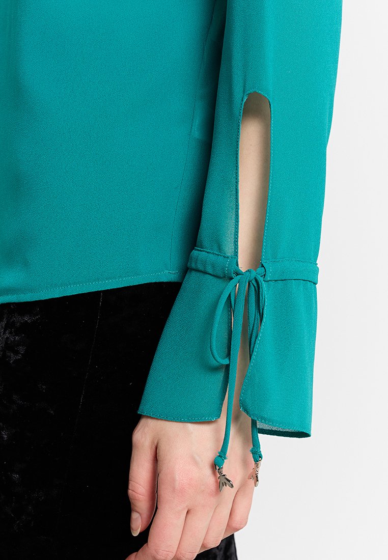 Türkises Langarmshirt mit Schlitz an den Handgelenken und gebundenen Kordeln, die mit metallischen Blattanhängern verziert sind, über einem schwarzen Samtrock.