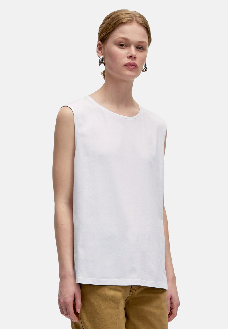 Un haut sans manches blanc en coton lisse, avec un col rond et une coupe simple. Associé à un pantalon beige. Le mannequin porte des boucles d'oreilles.