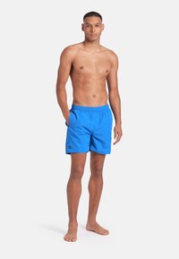 Giovane uomo in piedi a piedi nudi, indossa dei vivaci pantaloni da bagno blu, con una mano in tasca, su uno sfondo bianco uniforme.