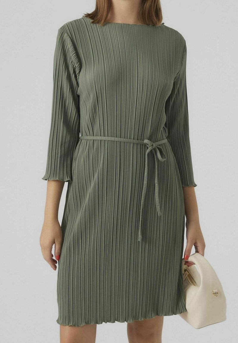 Femme portant une robe plissée longueur genou vert olive avec manches trois-quarts et ceinture à la taille, tenant un petit sac à main beige.