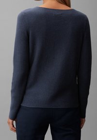 Osoba ubrana w ciemnoniebieski sweter z dzianiny z długim rękawem i ciemne spodnie, przedstawiona z tyłu na neutralnym szarym tle.