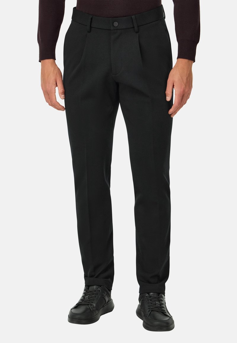 Homme portant un pantalon plissé noir, une ceinture noire, des baskets noires et un haut à manches longues sombre, debout devant un fond blanc.