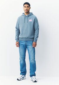 Šviesiai mėlynas džemperis su gobtuvu, priekyje kengūros tipo kišenė ir rožinės spalvos atspaustas logotipas, derinamas su šviesiai mėlynomis džinsinėmis kelnėmis ir baltomis sportinėmis avalynėmis su mėlynais akcentais.