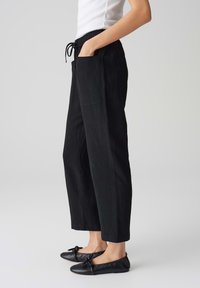 Pantalon noir à jambes larges en tissu doux, avec une taille élastique munie d'un cordon de serrage et des poches latérales. Porté avec des chaussures plates noires.