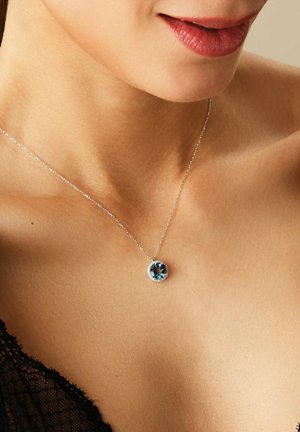 Silberne Halskette mit einem runden blauen Edelsteinanhänger, umgeben von kleinen klaren Steinen, die auf einer glatten Hautoberfläche liegen.