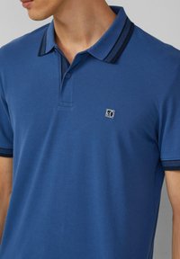 Niebieska koszulka polo wykonana z bawełny, z dwukolorowym kołnierzykiem w paski i małym logo na lewej piersi. Krótkie rękawy, teksturowana tkanina.