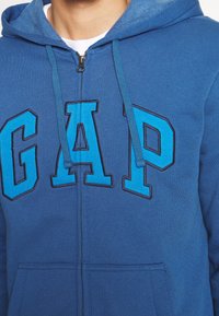 Sweat à capuche zippé bleu avec de grandes lettres "GAP" bleu clair sur la poitrine et des cordons assortis, porté par-dessus une chemise blanche.