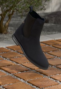Sort ankelstøvle med elastisk sidepanel svævende over firkantede træfliser med småsten i en udendørs setting.
