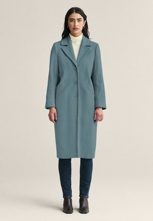 Kvinde står oprejst iført en midterlang teal-knappet frakke, hvid rullekrave, mørke jeans og brune ankelstøvler mod en ensfarvet baggrund.