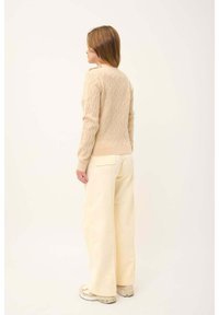 Maglione beige a maglia grossa con polsini a coste e un piccolo bottone sulla spalla, abbinato a pantaloni gialli chiari a gamba larga.