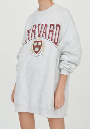Osoba nosząca oversize jasno szarą sukienkę z bluzy z czerwonym napisem Harvard i herbem z przodu, rękawy lekko podwinięte.