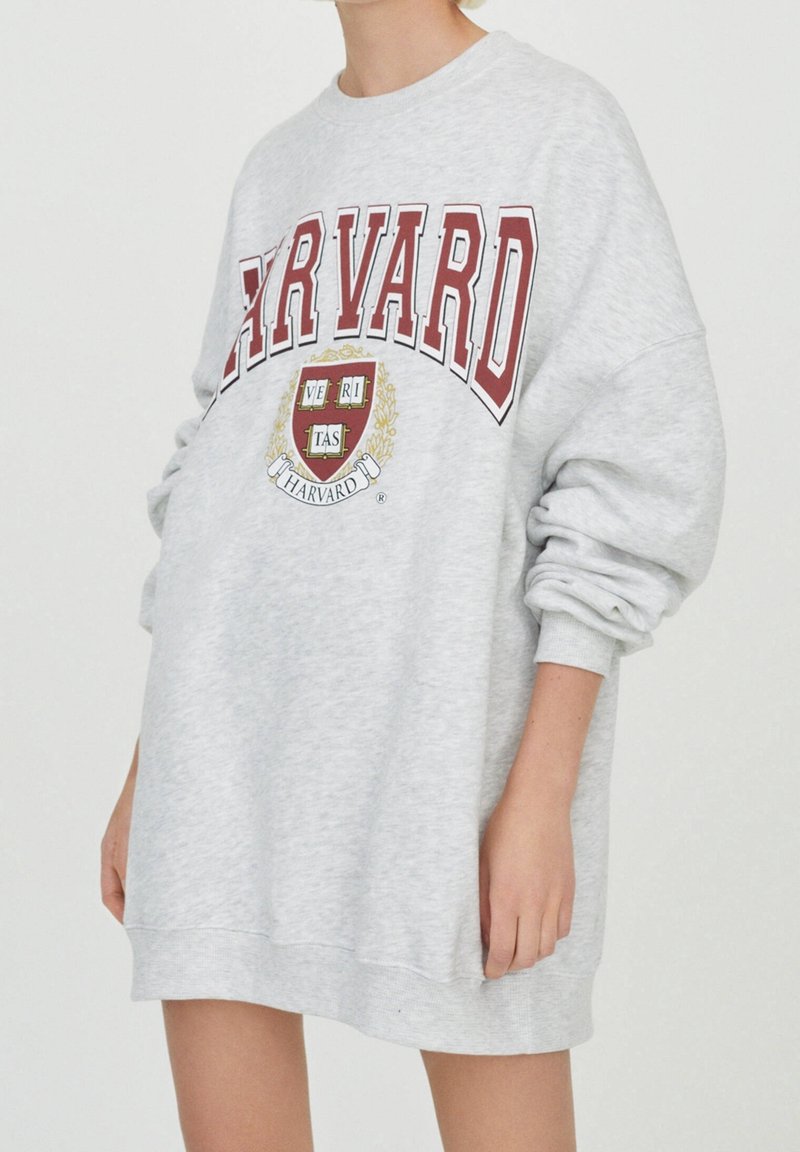 Personne portant une robe sweat-shirt gris clair oversize avec texte Harvard rouge et logo blason à l'avant, manches légèrement retroussées.