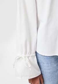 Weiße Bluse mit langen Ärmeln und gerafften Manschetten, die mit Kordelzügen ausgestattet ist. Glattes Gewebe mit weicher Textur, kombiniert mit hellblauen Jeans.