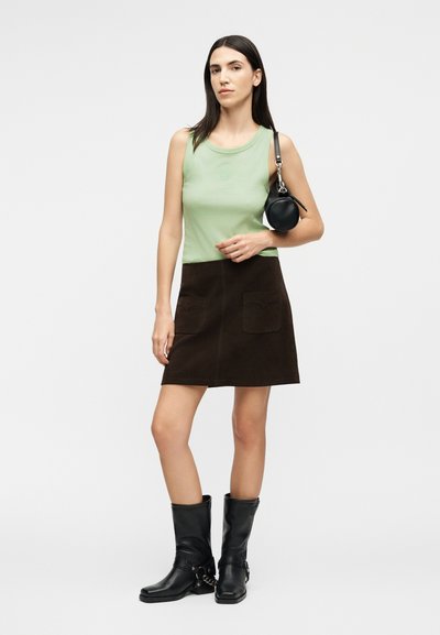 Γυναίκα με αμάνικο τοπ ανοιχτού πράσινου χρώματος, σκούρα καφέ φούστα με τσέπες, μαύρες μπότες μέχρι τη μέση της γάμπας, κρατώντας μικρή στρογγυλή μαύρη τσάντα στον ώμο.
