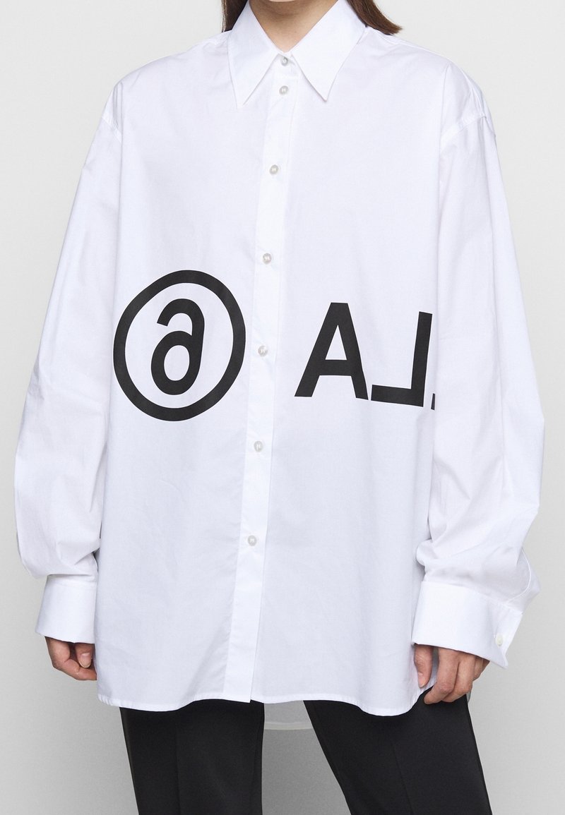 Vit oversized skjorta med knappar och spridd krage, med en stor svart logotyp och text framtill. Slät tygtextur.