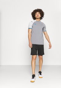 Grå sports t-skjorte med korte ermer, med blå detaljer og logo. Kombinert med svarte shorts og hvite sneakers med oransje såler.