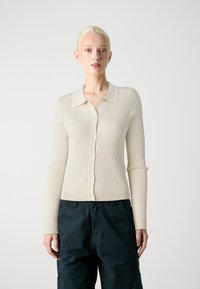 Ribbed, krémszínű kardigán, polo gallérral, hosszú ujjakkal és elől gombokkal. Slim fit fazonnal és texturált anyaggal rendelkezik.