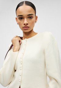 Balts vilnas kardigans ar apaļu kakla izgriezumu, priekšpuses aizdari ar dekoratīvām pogām un garām piedurknēm. Gluda tekstūra un pieguļošs dizains.