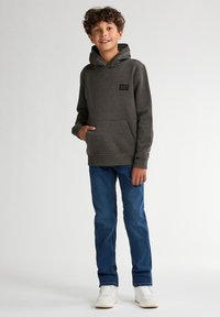 Felpa grigia con tasca frontale e cappuccio, abbinata a jeans blu e sneakers bianche. La felpa ha una piccola etichetta del marchio sul petto.