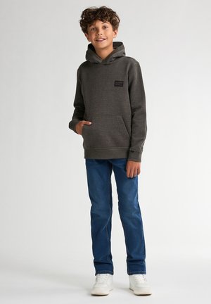 Felpa grigia con tasca frontale e cappuccio, abbinata a jeans blu e sneakers bianche. La felpa ha una piccola etichetta del marchio sul petto.