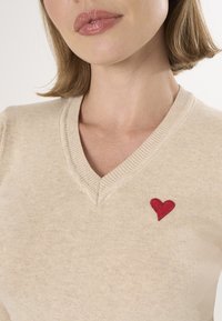 Beige v-hals trui gemaakt van zachte breiselstof, met een kleine geborduurde rode hart op de linkerzijde.