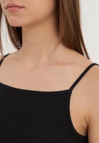 Débardeur noir à côtes avec fines bretelles, doté d'un décolleté carré et d'une texture douce, révélant une partie de l'épaule et du décolleté.