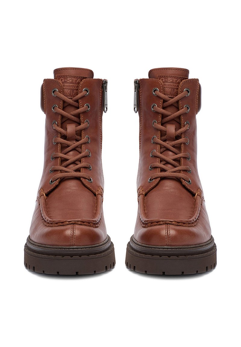 Un paio di stivali in pelle marrone con lacci, suola spessa scura e cerniere laterali, visti frontalmente su uno sfondo bianco.