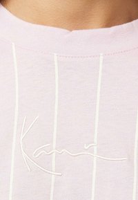 Svetloružové tričko s bielymi vertikálnymi pruhmi, zdobené vyšívaným podpisovým detailom. Jemný bavlnený materiál, okrúhly výstrih.