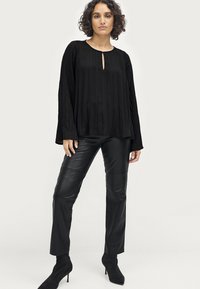 Schwarzes, plissiertes Langarmoberteil mit Schlüsselloch-Ausschnitt, kombiniert mit figurbetonten schwarzen Lederhosen und schwarzen High Heels.