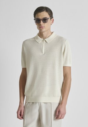 Uomo che indossa una polo a maniche corte beige in rete, pantaloni chiari e occhiali da sole scuri, in piedi davanti a uno sfondo grigio uniforme.