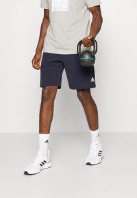 Marina atletiska shorts, en grå t-shirt med grafik, vita strumpor och vita sneakers med svarta accenter; håller en svart kettlebell.