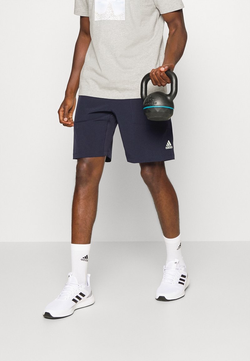 Marina atletiska shorts, en grå t-shirt med grafik, vita strumpor och vita sneakers med svarta accenter; håller en svart kettlebell.