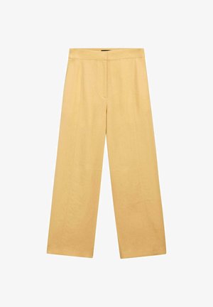 Kobieta w luźnym, żółtym, bezrękawnym żakiecie i pasujących szerokich spodniach, stojąca na tle białej ściany, ręce w kieszeniach.