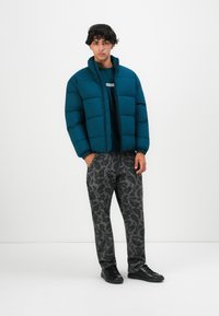Joven vestido con una chaqueta acolchada verde azulado, suéter verde azulado, pantalones grises y negros con estampado, y zapatos negros, de pie frente a un fondo blanco liso.
