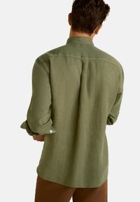Olivenfarbendes Leinenhemd mit lockerer Passform, aufgerollten Ärmeln und einem kleinen Kragen. Es hat eine mittige Rückennaht und weiße Knöpfe.