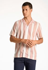 Camicetta a maniche corte con bottoni, a righe verticali rosa e bianche. Tessuto leggero e colletto da campeggio. Indossata con pantaloni blu scuro.
