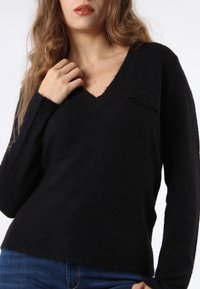 Maglione nero realizzato in materiale morbido, con scollo a V, orli a coste e un piccolo logo sul petto. Maniche lunghe, vestibilità comoda.